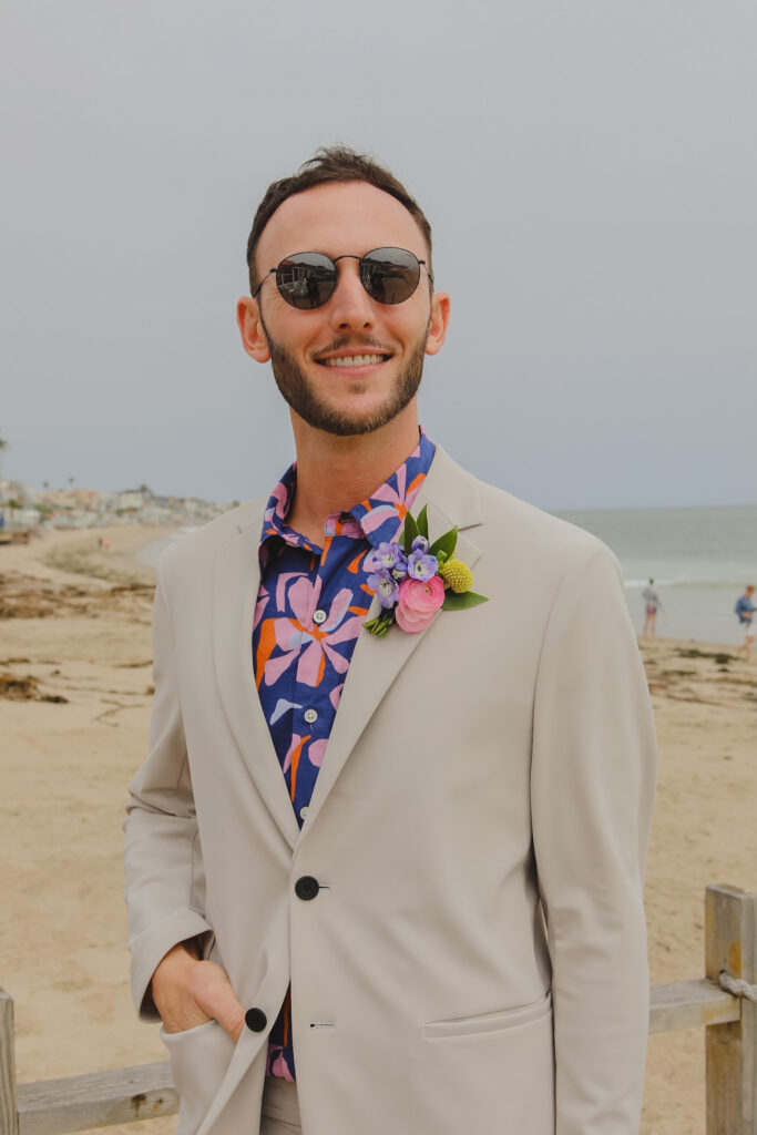 groom in bold print shirt and tan suit with colorful boutonniere stands on beach for private beach wedding ceremony