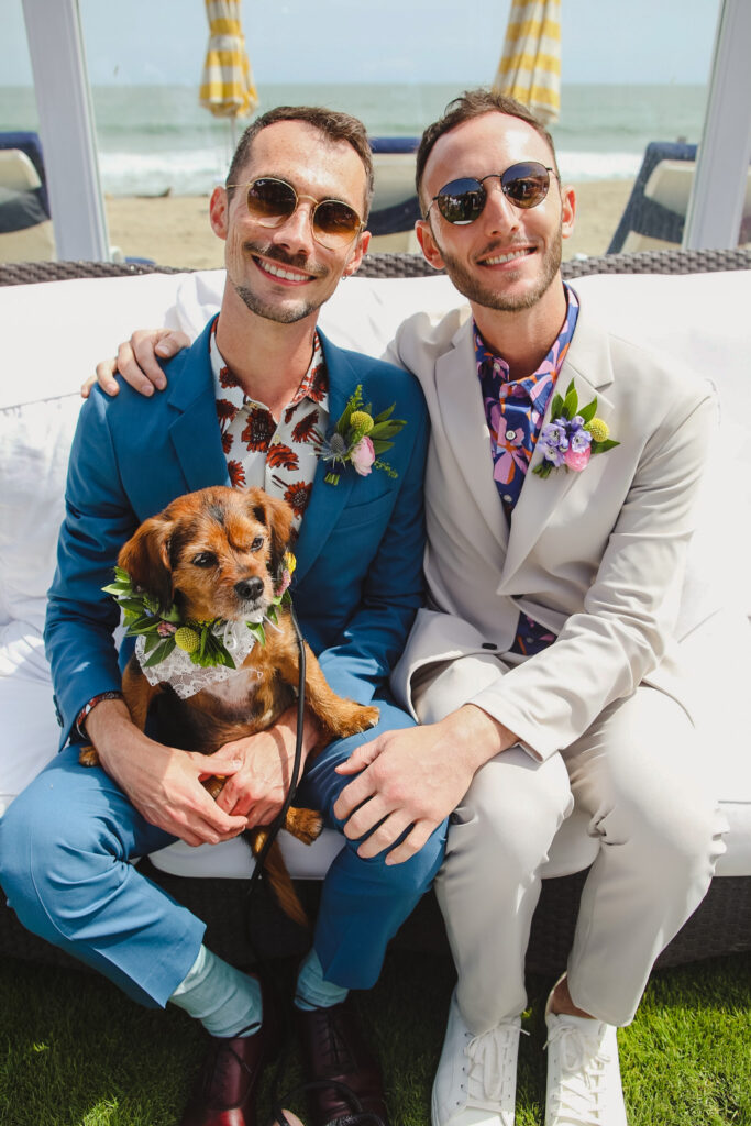 groom in bold print shirt and blue suit and groom in bold print shirt with tan suit hold dog with flower collar 