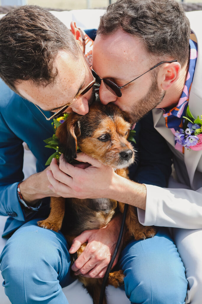 groom in bold print shirt and blue suit and groom in bold print shirt with tan suit kiss dog with flower collar 