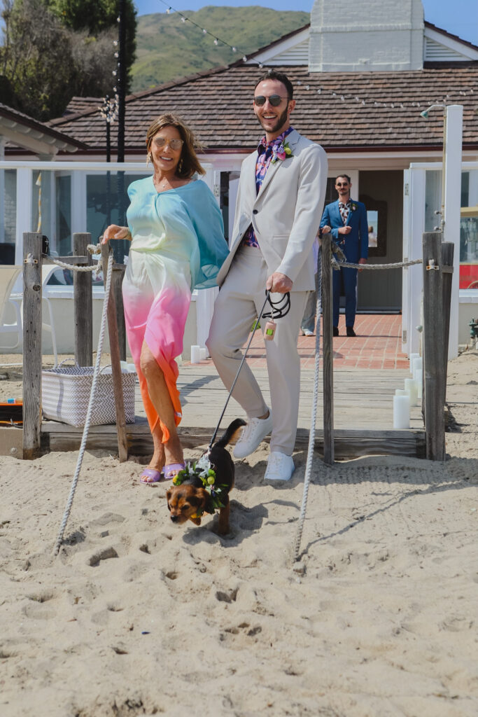groom in bold print shirt and tan suit walks with mother and dog with flower collar down wedding ceremony aisle