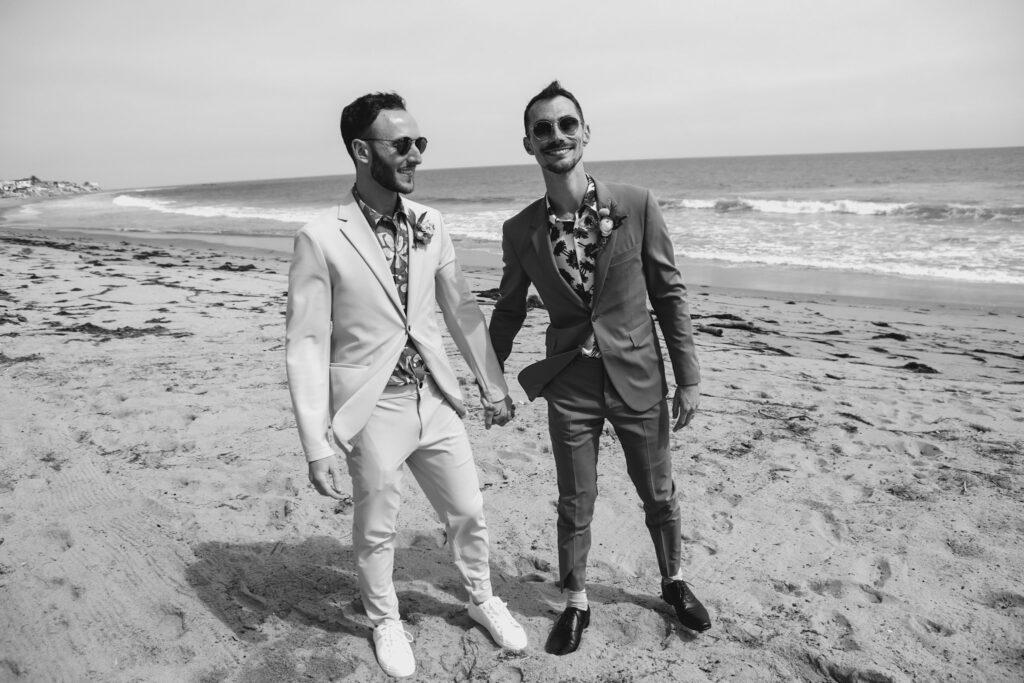 groom in bold print shirt and tan suit walks with groom in bold print shirt and blue suit on the beach