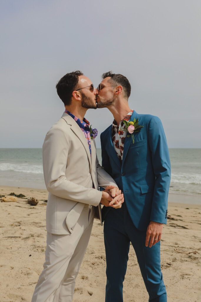 groom in bold print shirt and tan suit and groom in bold print shirt and blue suit kiss on the beach