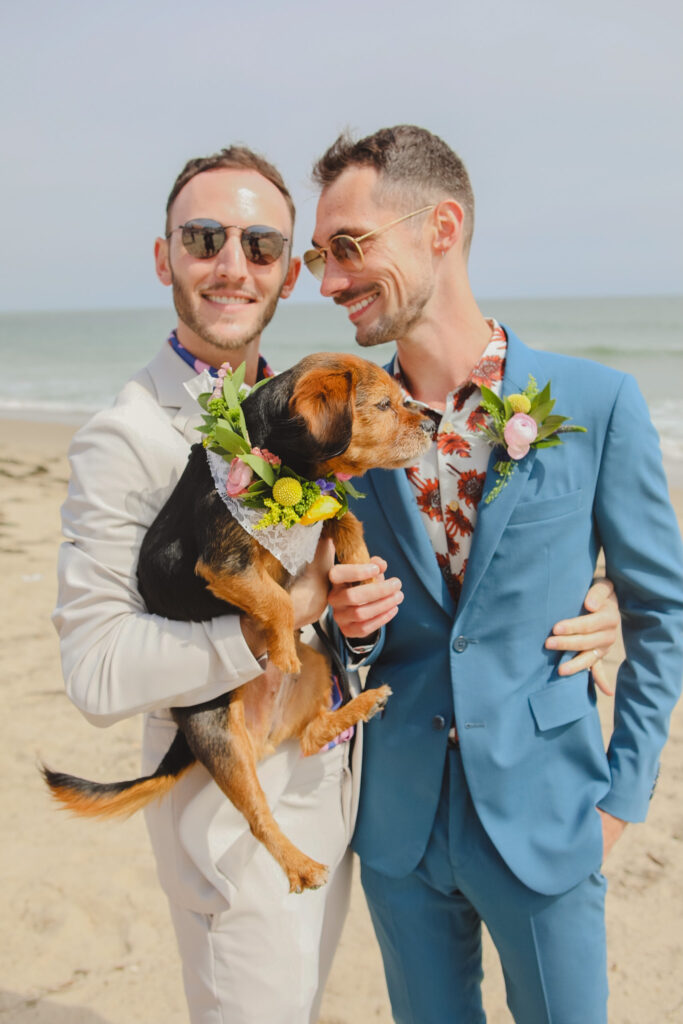 groom in tan suit and groom in blue suit hold dog with flower collar on the beach