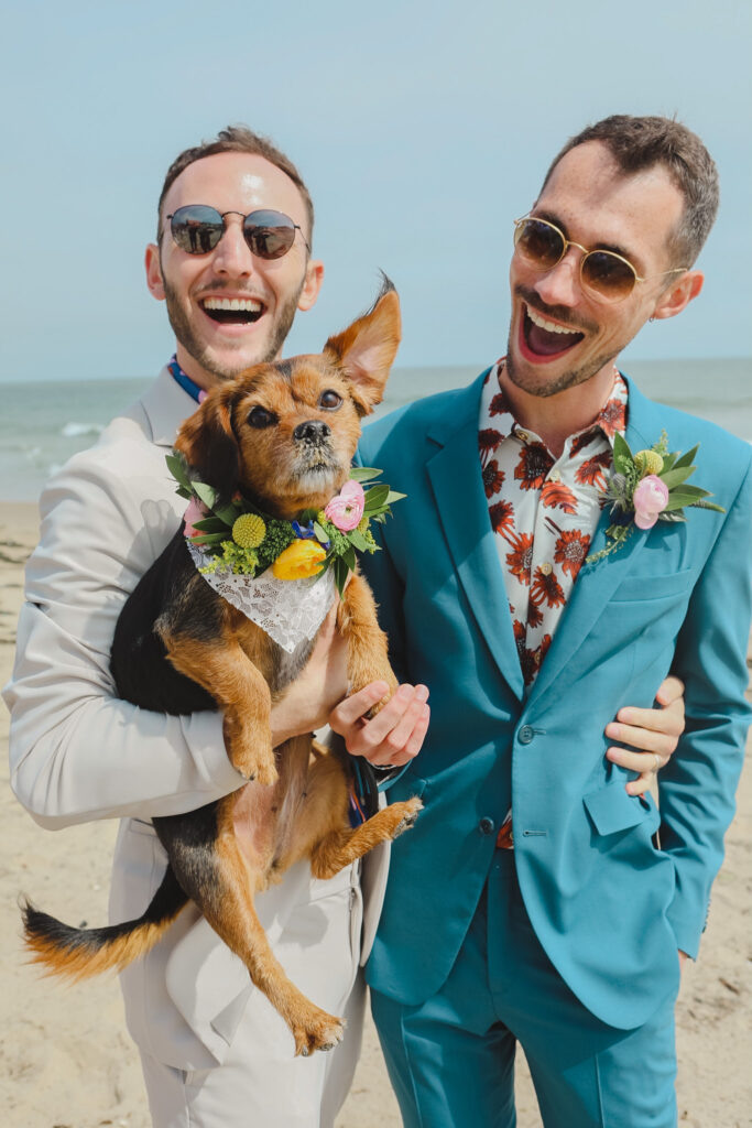 groom in tan suit and groom in blue suit hold dog with flower collar on the beach
