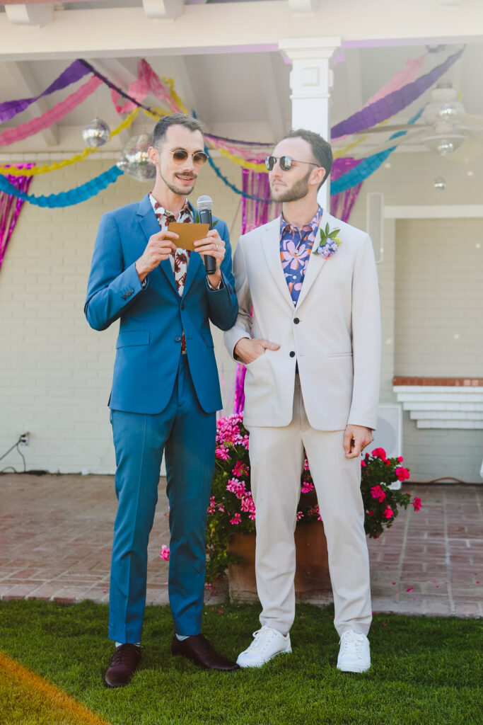 groom in tan suit and groom in blue suit give speech to guests 