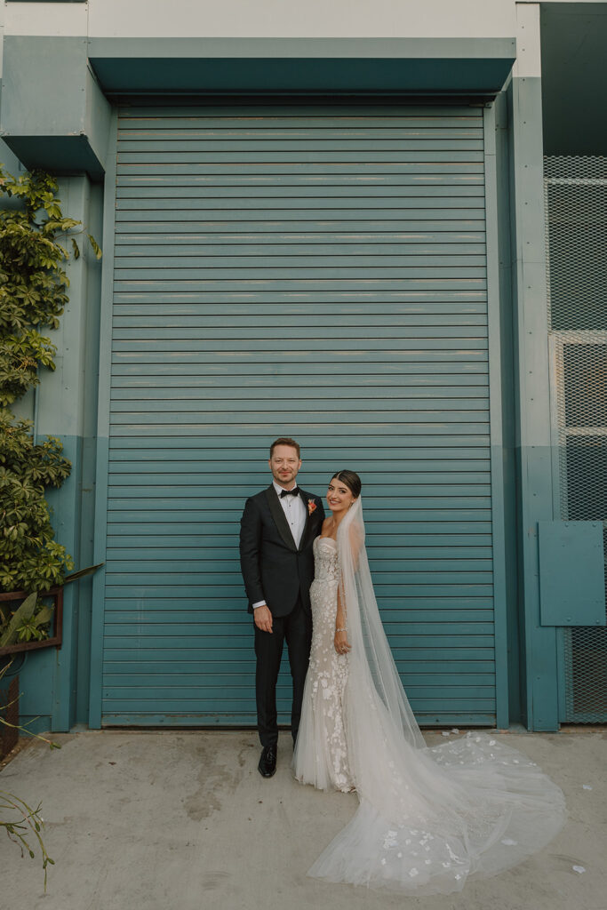 bride in strapless wedding dress with 3d floral appliqué stands with groom in black tuxedo in front of green wall at Valentine DTLA