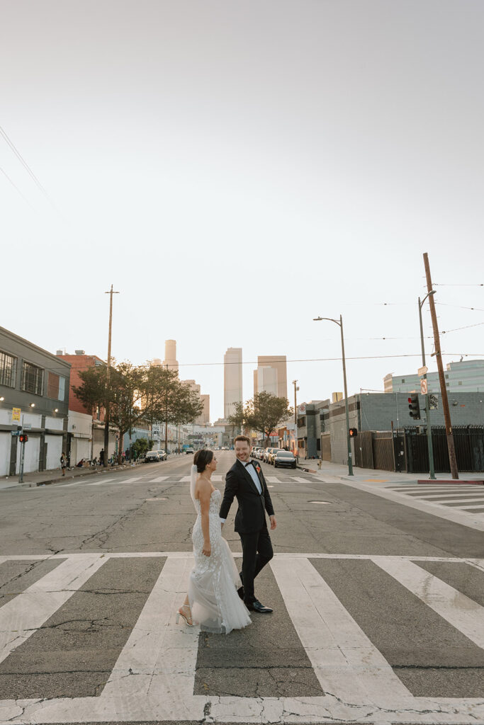 bride in strapless wedding dress with 3d floral appliqué and pink tropical bridal bouquet walks with groom in black tuxedo in DTLA
