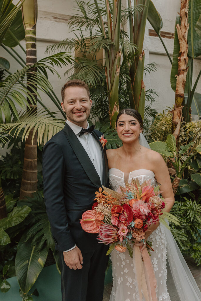 bride in strapless wedding dress with 3d floral appliqué and pink tropical bridal bouquet stands with groom in black tuxedo