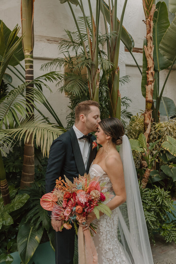 bride in strapless wedding dress with 3d floral appliqué and pink tropical bridal bouquet stands with groom in black tuxedo
