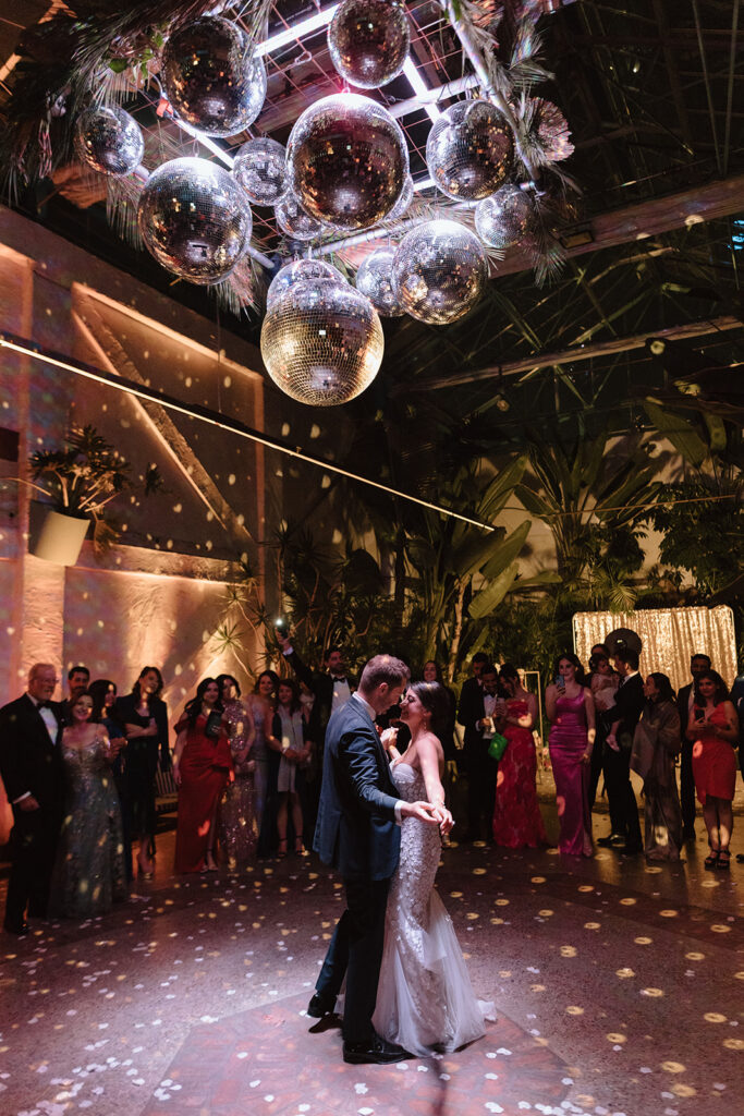 bride in strapless wedding dress with 3d floral appliqué and groom in black tuxedo have first dance under large disco ball installation at Valentine