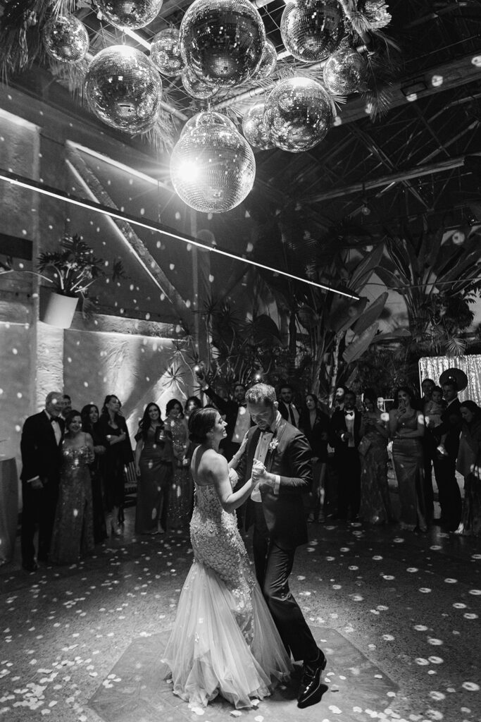 bride in strapless wedding dress with 3d floral appliqué and groom in black tuxedo have first dance under large disco ball installation at Valentine