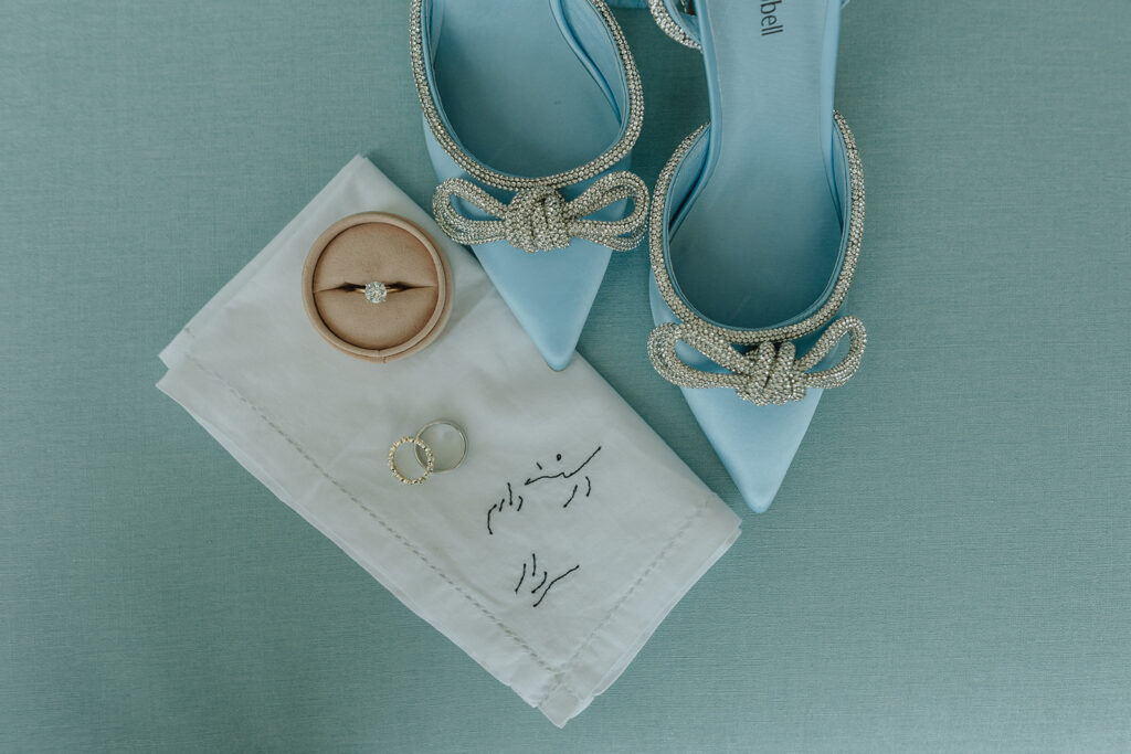 something blue bridal shoes with crystal bows next to customized handkerchief and rings