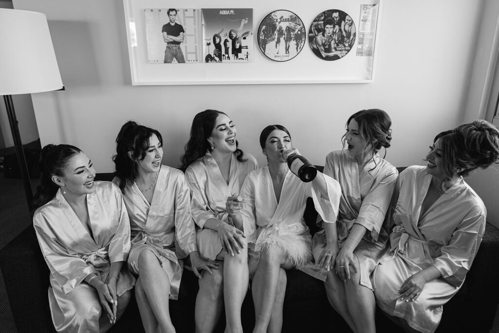 bride in white satin robe drinking champagne with bridesmaids in satin robes