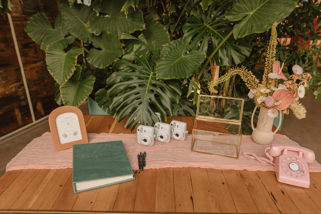 wedding welcome table at Valentine DTLA with pink phone, gold glass card box, dark green guest book and white polaroid cameras