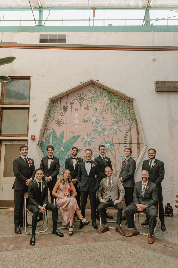 groom in black tuxedo stands with co-ed wedding party in mixed suits in front of pink keyhole wall at Valentine DTLA