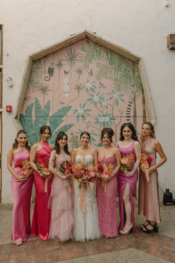 bride in strapless wedding dress with 3d floral appliqué standing with bridesmaids in mix matched pink Barbiecore dresses in front of pink keyhole wall at Valentine DTLA