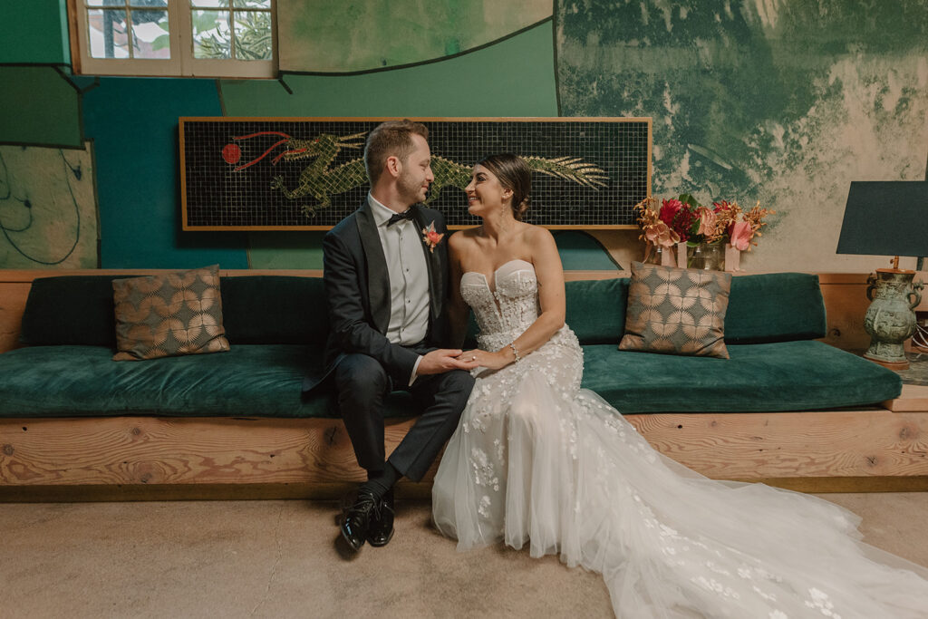 bride in strapless wedding dress with 3d floral appliqué sits with groom in black tuxedo in green art room at Valentine DTLA