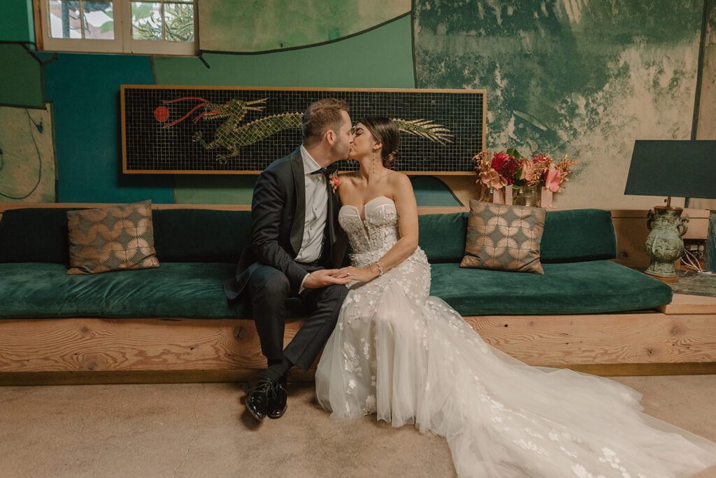 bride in strapless wedding dress with 3d floral appliqué kisses groom in black tuxedo in green art room at Valentine DTLA