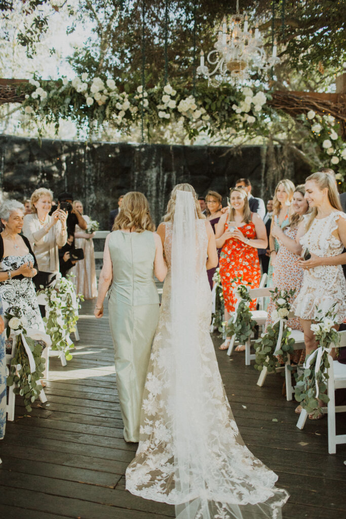 bride in lace dress with cathedral length veil walks down ceremony aisle with mother in green dress for waterfall wedding ceremony at The Oak Room at Calamigos Ranch