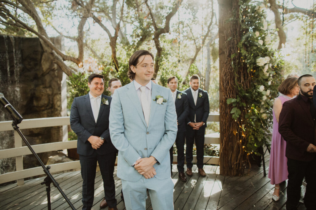 groom in light blue suit watching partner walk down ceremony aisle with groomsmen in dark grey suits behind him