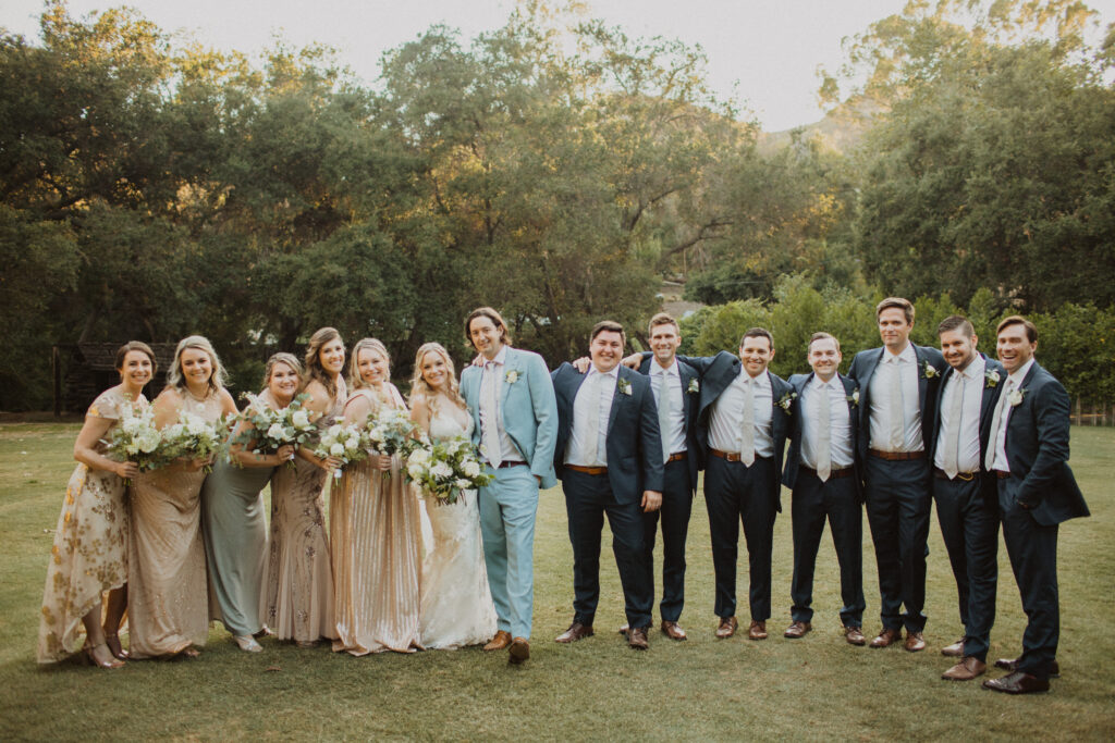 groom in light blue suit and bride in lace wedding dress stand with wedding party in dark grey suits and mix matched soft gold bridesmaid dresses