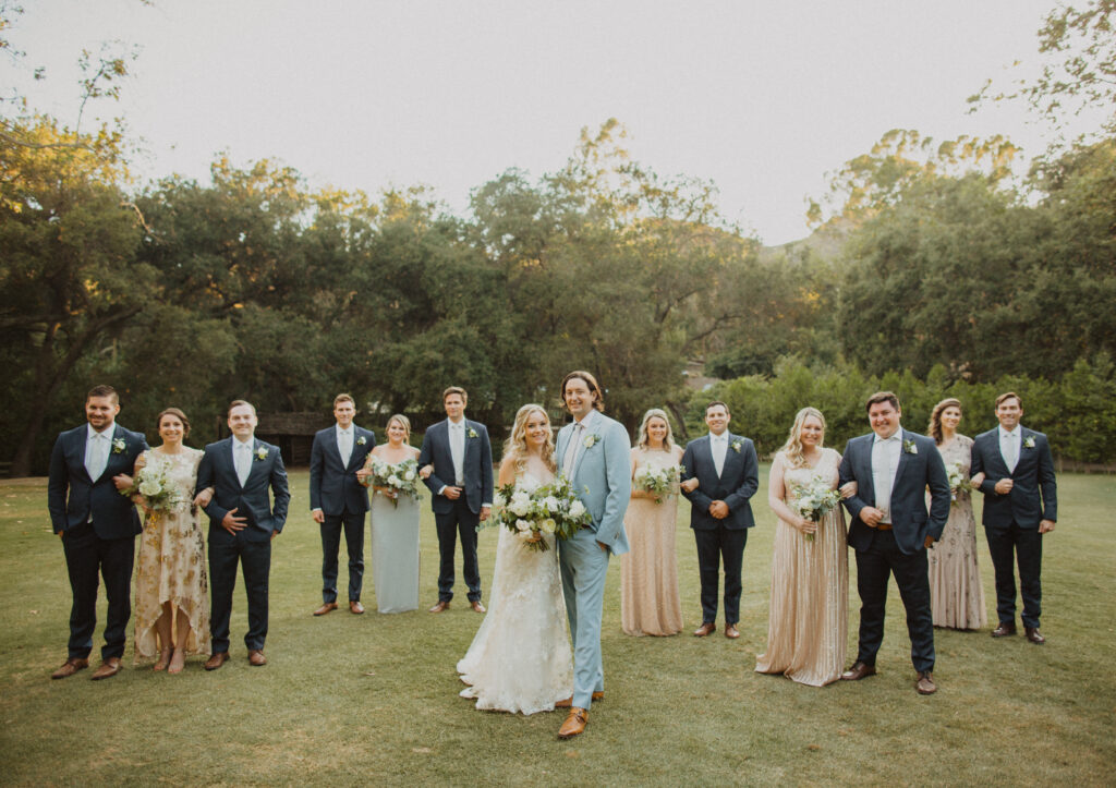 groom in light blue suit and bride in lace wedding dress stand with wedding party in dark grey suits and mix matched soft gold bridesmaid dresses