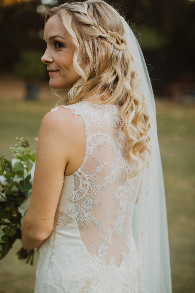 bride in lace wedding dress with embellished open back and  floor length veil holding white floral bouquet