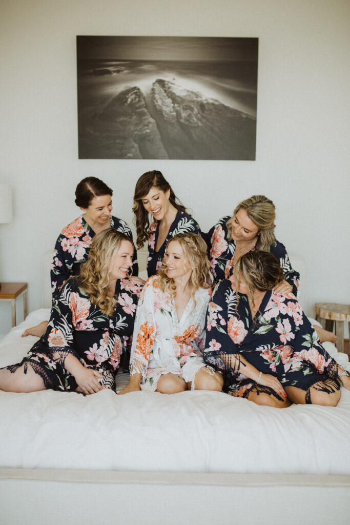 bride in white floral robe sitting with bridesmaids in dark floral robes on bed while getting ready for wedding