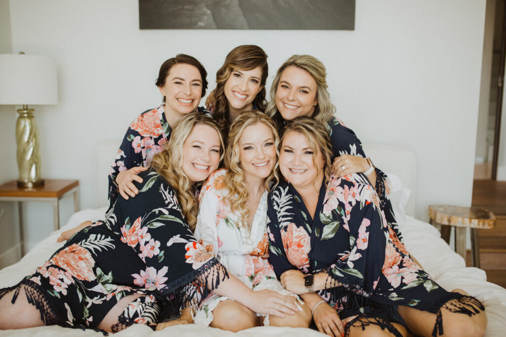 bride in white floral robe sitting with bridesmaids in dark floral robes on bed while getting ready for wedding
