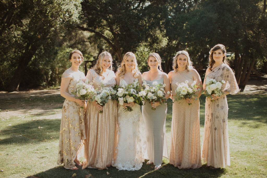 bride in lace wedding dress with bridesmaids in mix matched soft gold dresses all holding white floral bouquets
