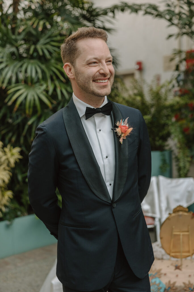 groom in black tuxedo with pink dried boutonniere waits at end of ceremony aisle