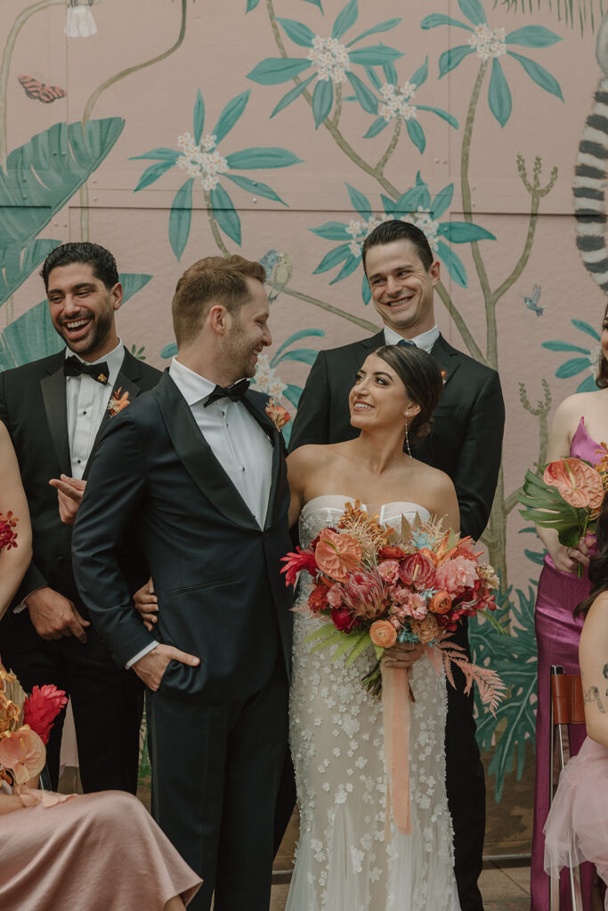 bride in strapless wedding dress with 3d floral appliqué holding tropical bouquet standing and groom in black tuxedo stands with wedding party in mix matched pink Barbiecore dresses and black suits in front of pink keyhole wall at Valentine DTLA