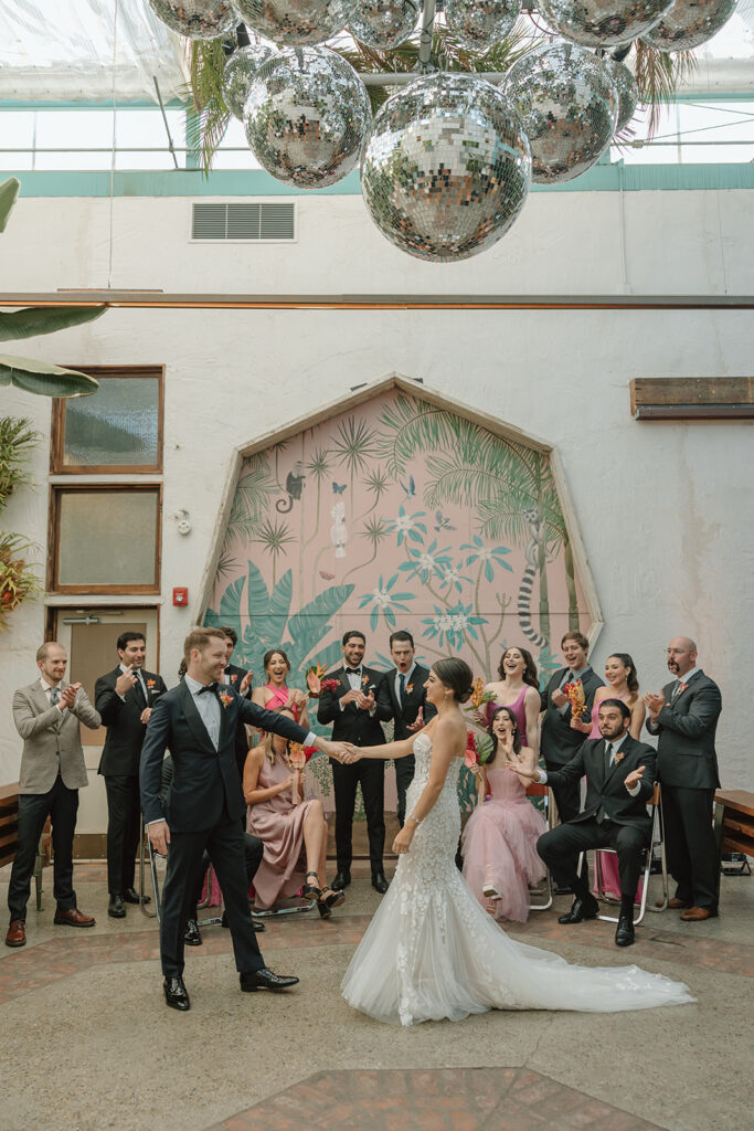 bride in strapless wedding dress with 3d floral appliqué holding tropical bouquet standing and groom in black tuxedo stands with wedding party in mix matched pink Barbiecore dresses and black suits in front of pink keyhole wall at Valentine DTLA