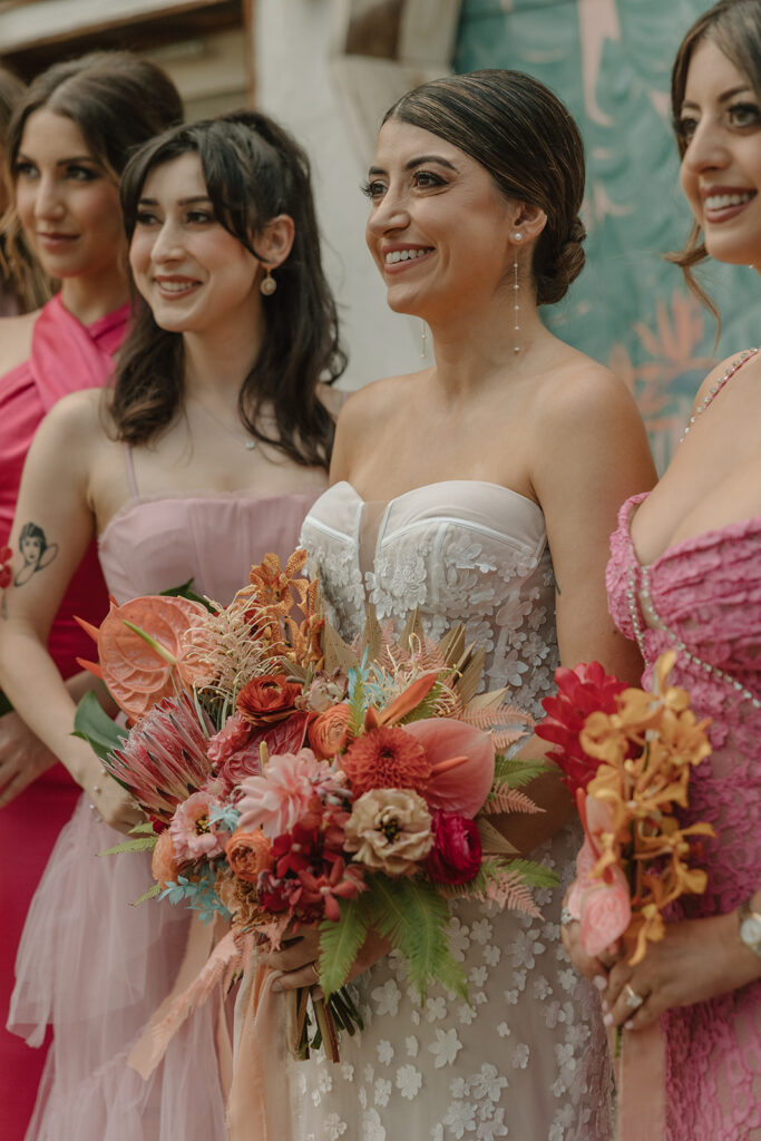 bride in strapless wedding dress with 3d floral appliqué holding tropical bouquet standing with bridesmaids in mix matched pink Barbiecore dresses in front of pink keyhole wall at Valentine DTLA