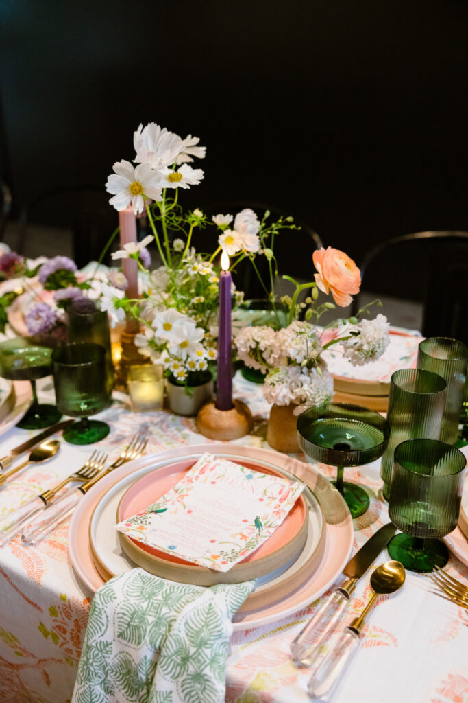 colorful summer dinner table decor with green glassware, taper candles,  wooden plates, floral table cloth and napkins and fresh floral centerpieces 
