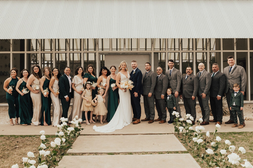bride in deep v-neck wedding dress with groom in dark navy suit standing with wedding party in dark grey suits, dark green and champagne outfits