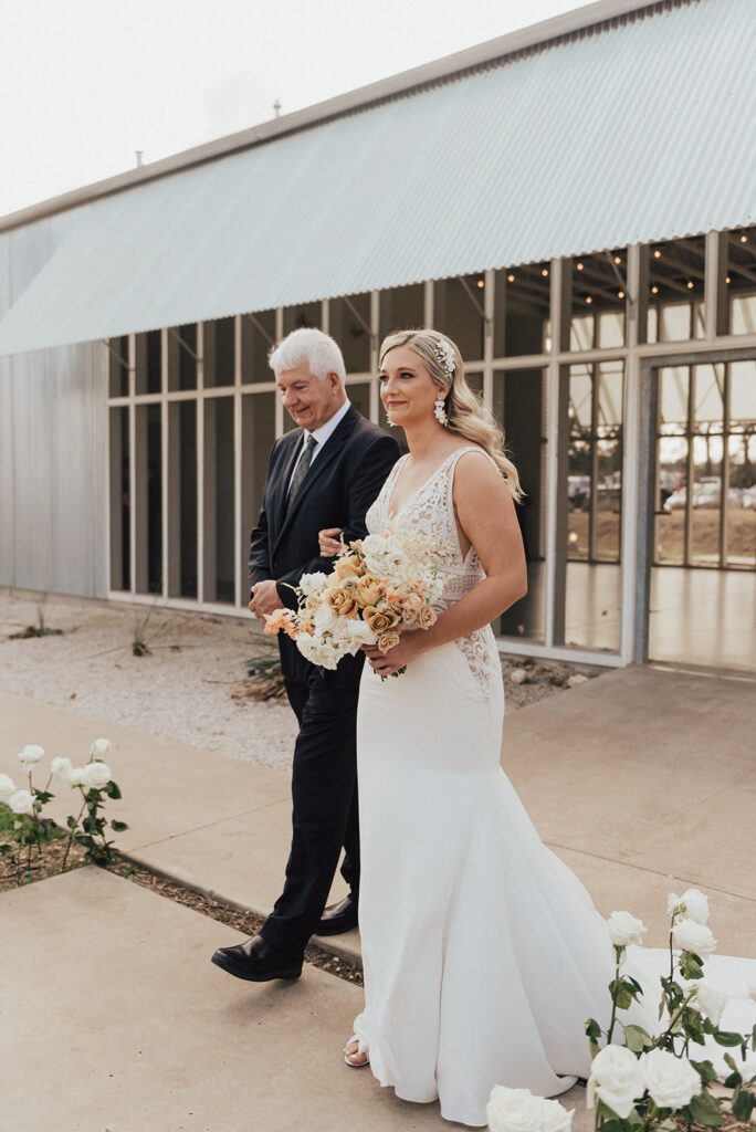 bride in deep v neck wedding dress and white floral bouquet walks with father down aisle
