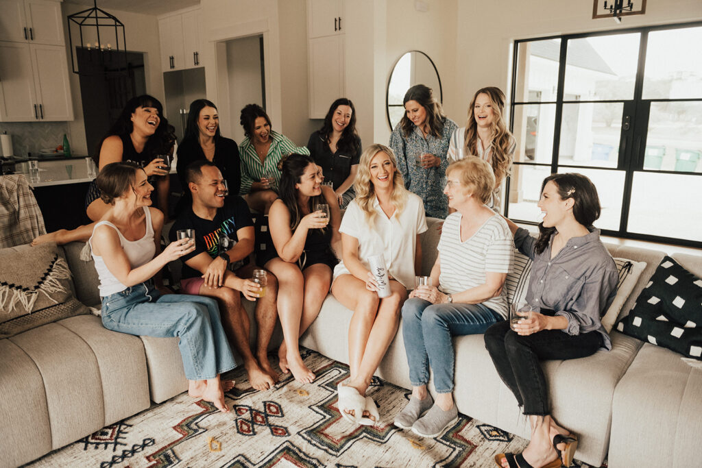 bride to be in satin pajamas sitting on couch with co-ed wedding party before wedding