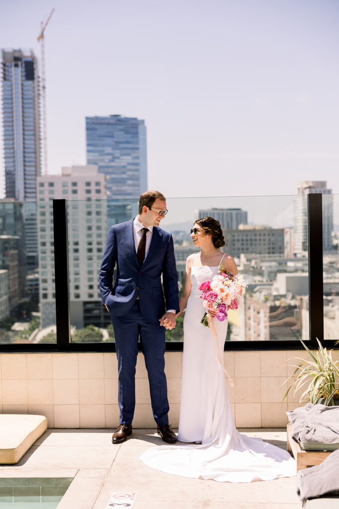 groom in dark blue suit and black tie stands with bride in minimalist wedding dress with DTLA skyline in background