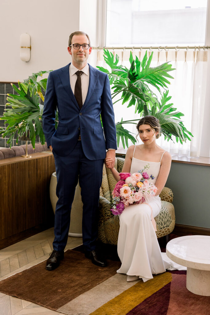groom in dark blue suit and black tie stands with bride in minimalist wedding dress and colorful bouquet