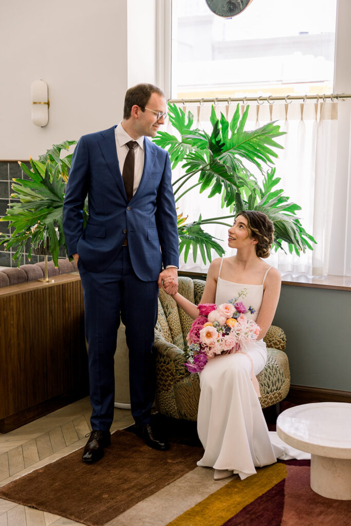 groom in dark blue suit and black tie stands with bride in minimalist wedding dress and colorful bouquet