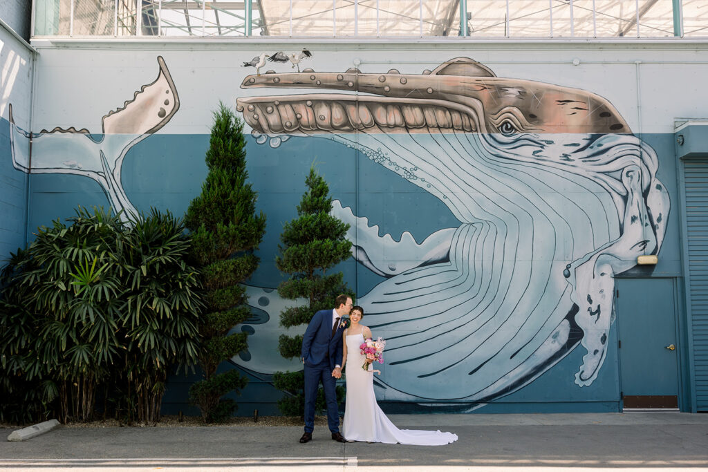 groom in dark blue suit and black tie stands with bride in minimalist wedding dress and colorful bouquet in front of whale mural at The Grass Room