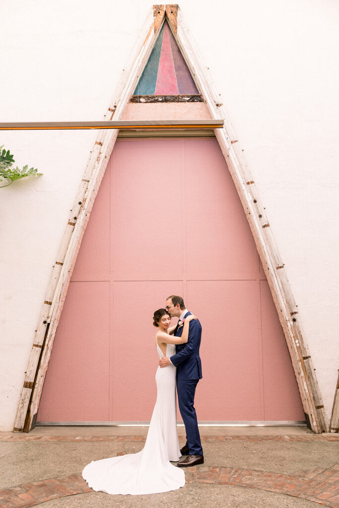 bride in minimalist wedding dress and groom in dark blue suit stand in front of pink keyhole wall at Grass Room DTLA