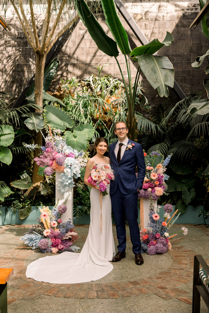 bride in minimalist wedding dress and groom in dark blue suit stand with pastel ceremony floral pillars surrounded by greenery at The Grass Room DTLA
