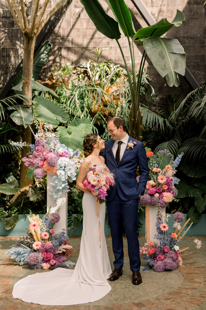 bride in minimalist wedding dress and groom in dark blue suit stand with pastel ceremony floral pillars surrounded by greenery at The Grass Room DTLA