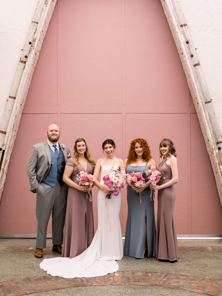 bride in modern wedding dress stands with wedding party in mix of pink, dark grey and blue outfits