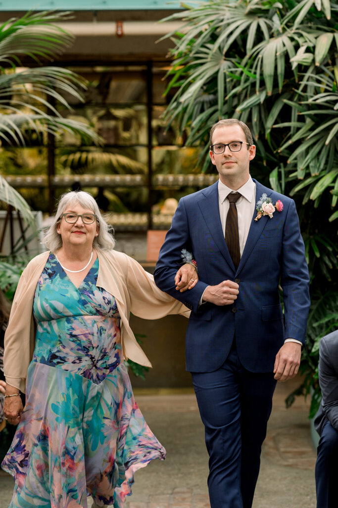 groom in glasses and dark blue suit walks ceremony aisle with mother 