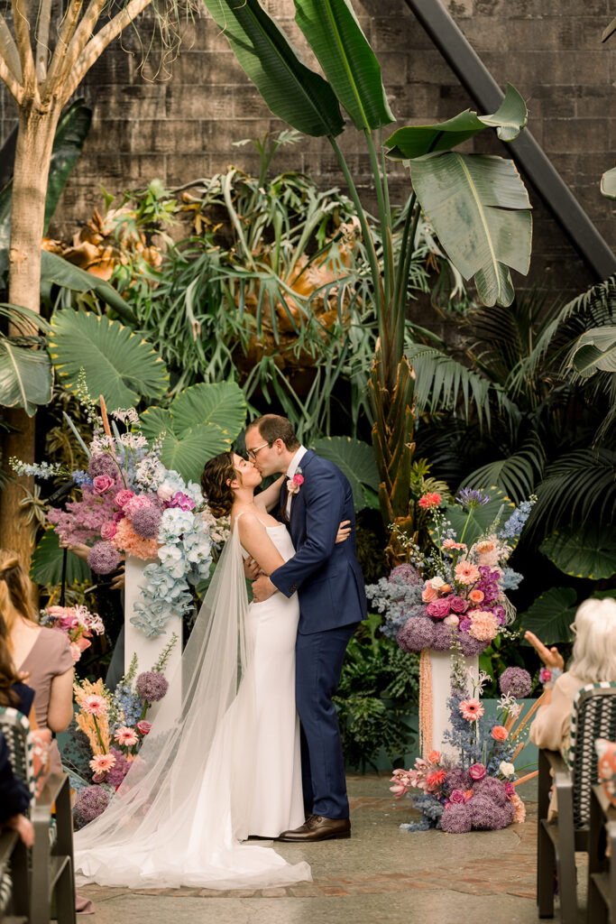 bride in modern wedding dress with bridal cape share first kiss with groom in dark blue suit during contemporary pastel wedding at The Grass Room DTLA