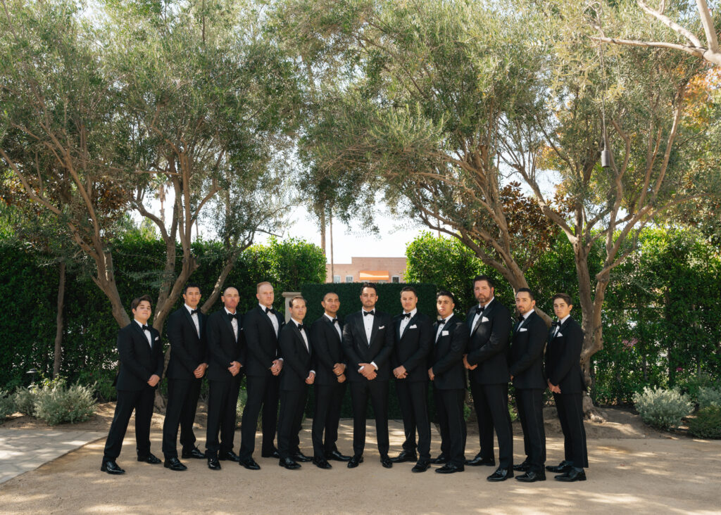groom in classic black tuxedo suit with bowtie stands with groomsmen in matching suits