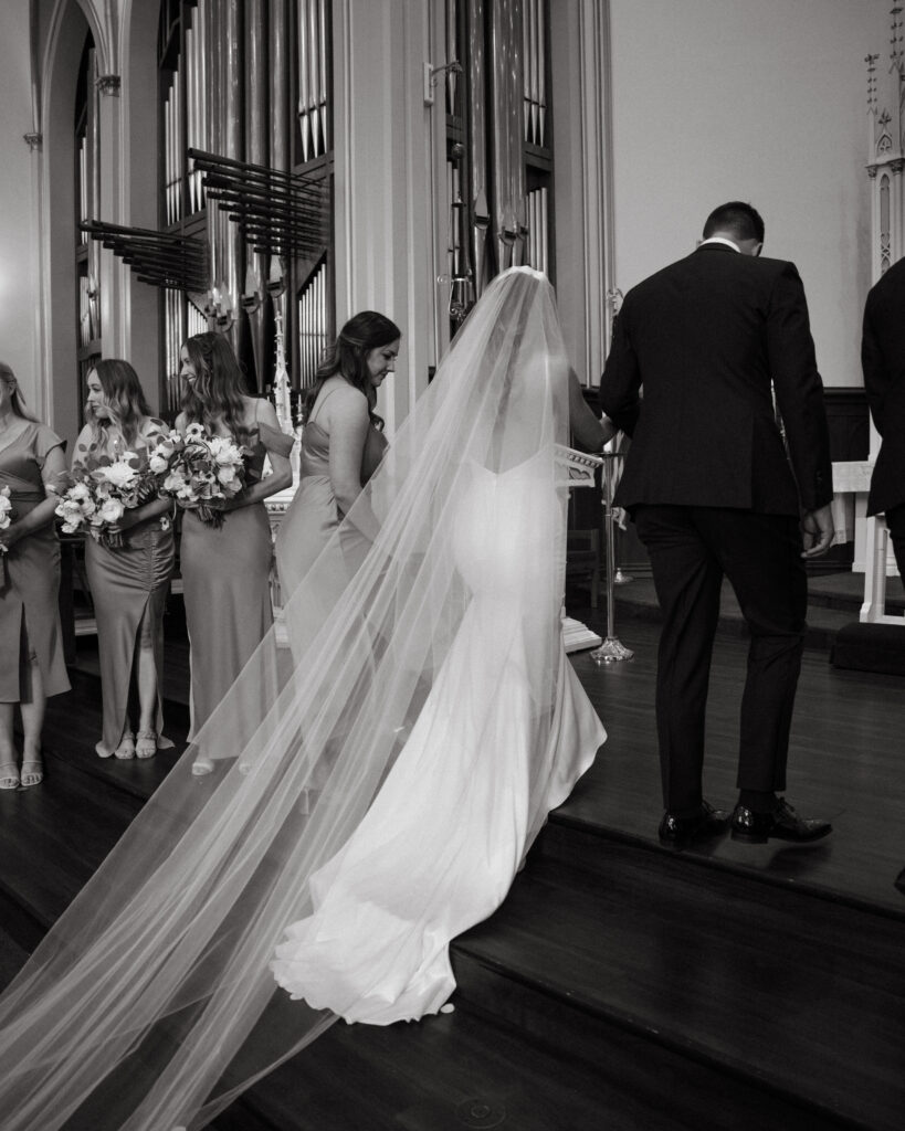 bride in cathedral veil stands with groom during church wedding ceremony